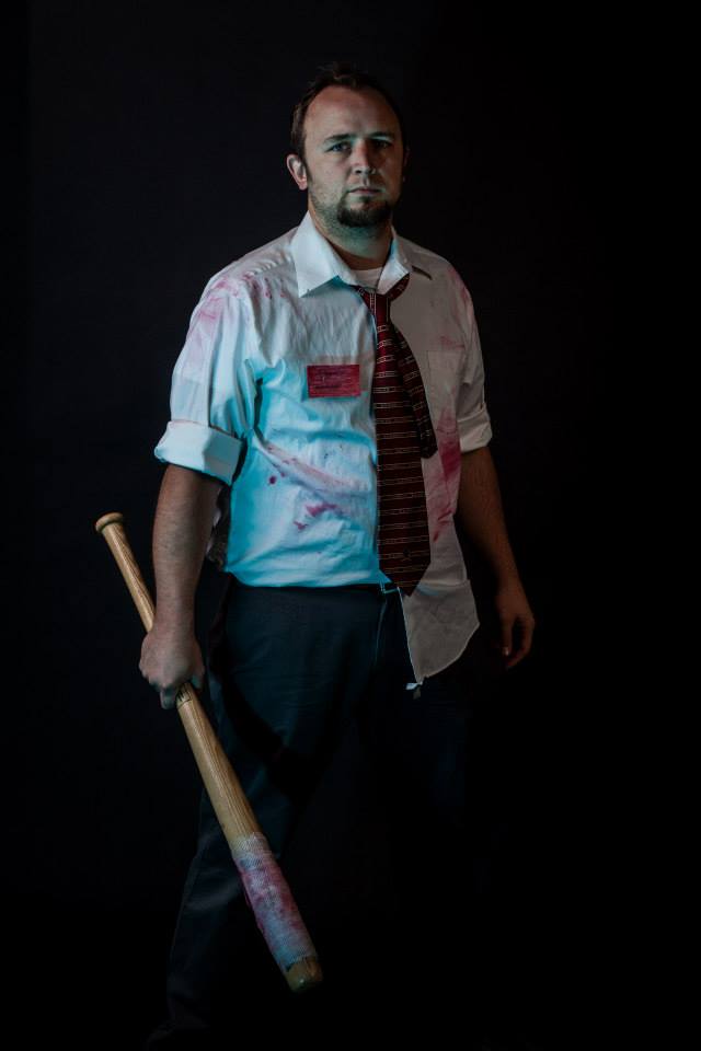 Image of Jeremy in a white shirt and striped tie stands holding a baseball bat, with red stains on his clothes, suggesting a horror theme